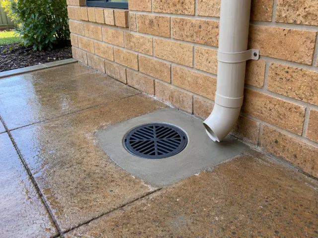 Outdoor gully grate beside wet brick paving and a downpipe on a brick wall in Raworth