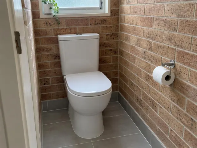 White toilet installed in a small brick-lined bathroom with tiled floor and window in Tenambit